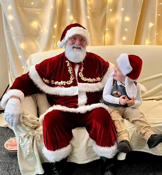 Santa Claus and a young boy sit on a cream colored coach in front of a cream background illuminated with Christmas lights.