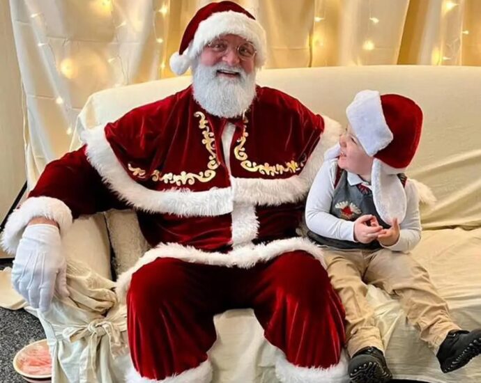 Santa Claus and a young boy sit on a cream colored coach in front of a cream background illuminated with Christmas lights.