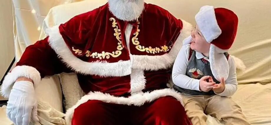Santa Claus and a young boy sit on a cream colored coach in front of a cream background illuminated with Christmas lights.